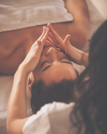 Atpalaiduojantis mioliftingo masažas veidui – moteris guli ant masažo stalo, uždengta rankšluosčiu, o specialistė švelniai masažuoja jos veidą. Procedūra skirta veido odos stangrinimui ir atjauninimui, skatinant kraujotaką ir kolageno gamybą.