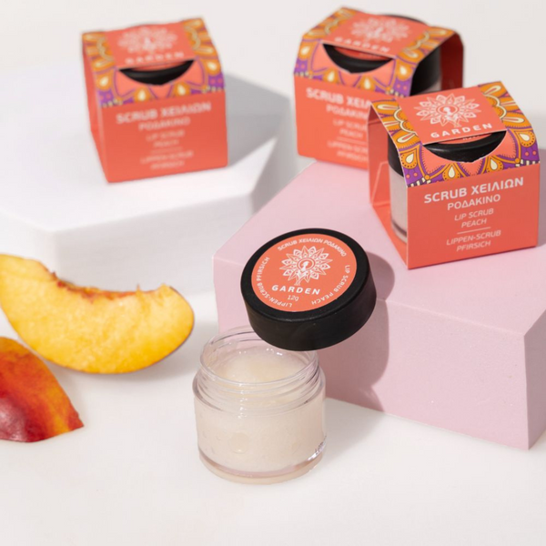 „GARDEN Peach lūpų šveitiklis stiklainėlyje su atidarytu dangteliu, cukrinė tekstūra, persiko skiltelės ir spalvinga pakuotė fone“